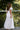 Woman in a white dress standing on a wooden deck with greenery in the background