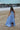 girl in a blue dress standing on a sandy beach with boats in the background