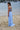 Girl in a light blue dress standing on a beach with water and boats in the background